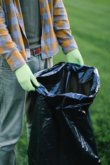 A street scene showing a white commercial rubbish collection truck with a rusty, open rear compactor area positioned near a sidewalk, actively engaged in waste collection. A worker dressed in a high-v