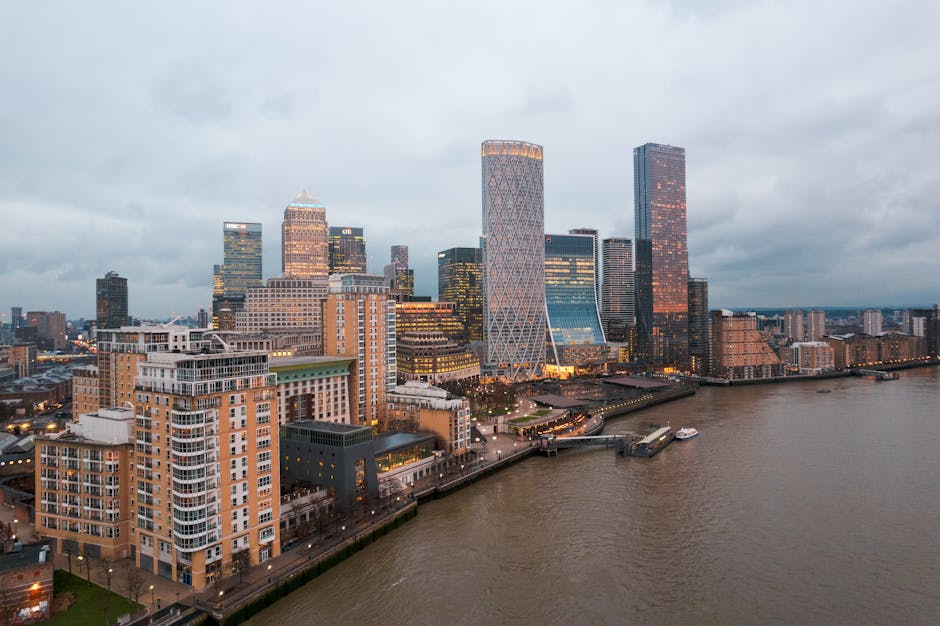 A black-and-white photograph showcasing a modern urban cityscape with multiple high-rise office buildings and commercial structures. In the foreground, a large, rounded, multi-storey building features